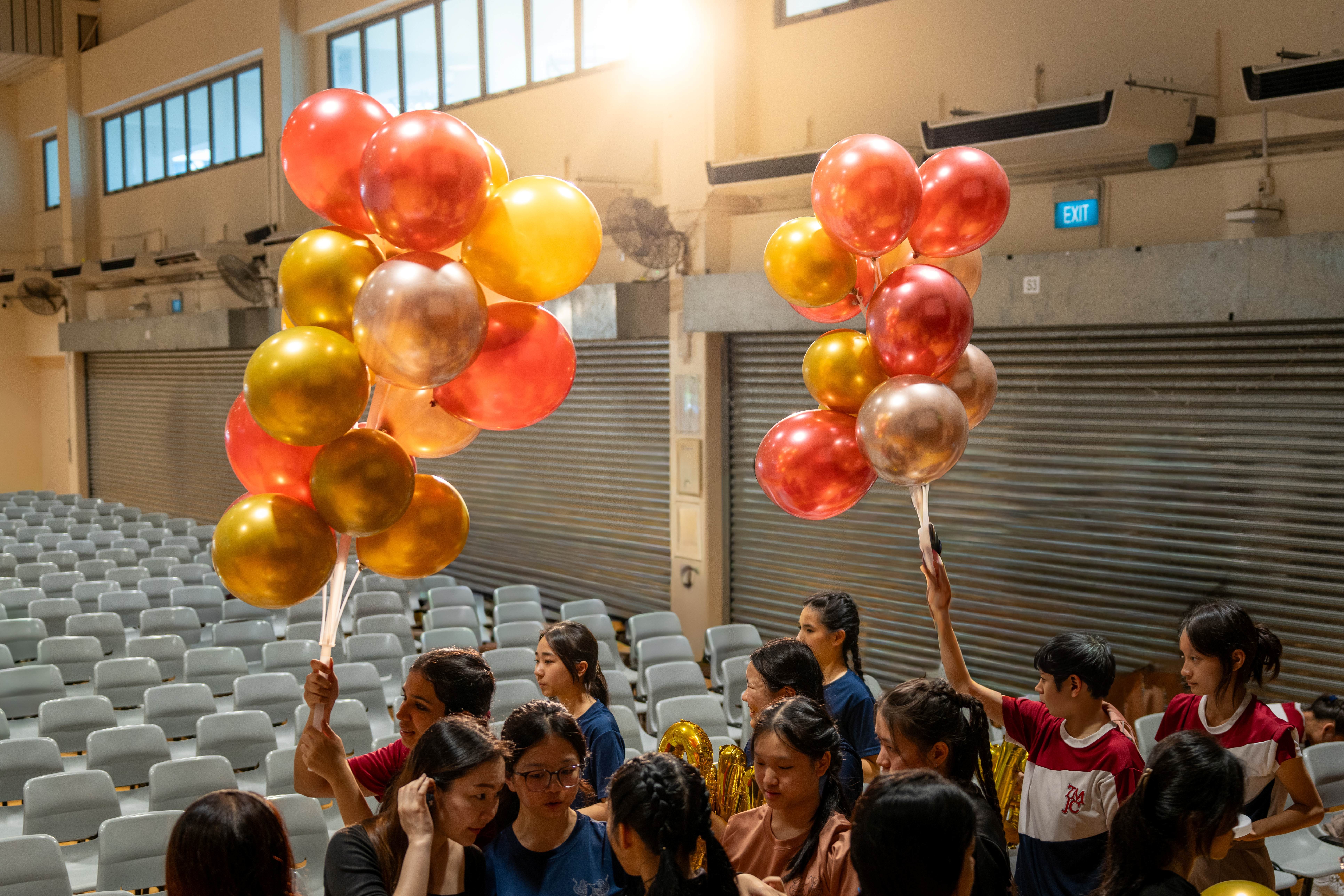 Students in a school hall hold up large clusters of red, gold, and rose-gold balloons while preparing for an event.